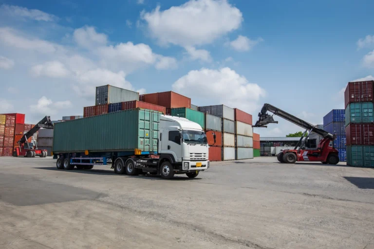 A white truck is positioned in a logistics yard surrounded by colorful shipping containers, showcasing HS Agency's expertise in transportation and warehousing solutions. The image highlights efficient supply chain management, essential for seamless domestic and international logistics services.